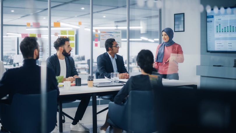 Picture of a board room with three men and a woman looking at a Muslim woman presenting who wears a rose colored sweater and a blue hijab.