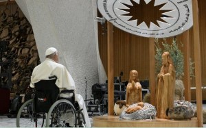 Pope Francis sits in a wheel chair before a large nativity.